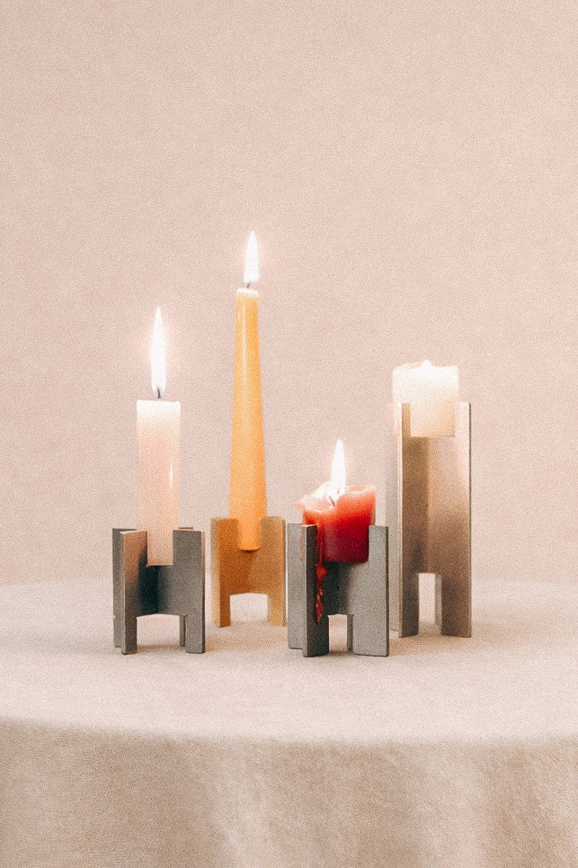 Minimalist Lyssier candleholders in brushed and polished metal, holding lit taper and pillar candles in different sizes, arranged on a neutral background showcasing material contrast, precision, and sculptural form.