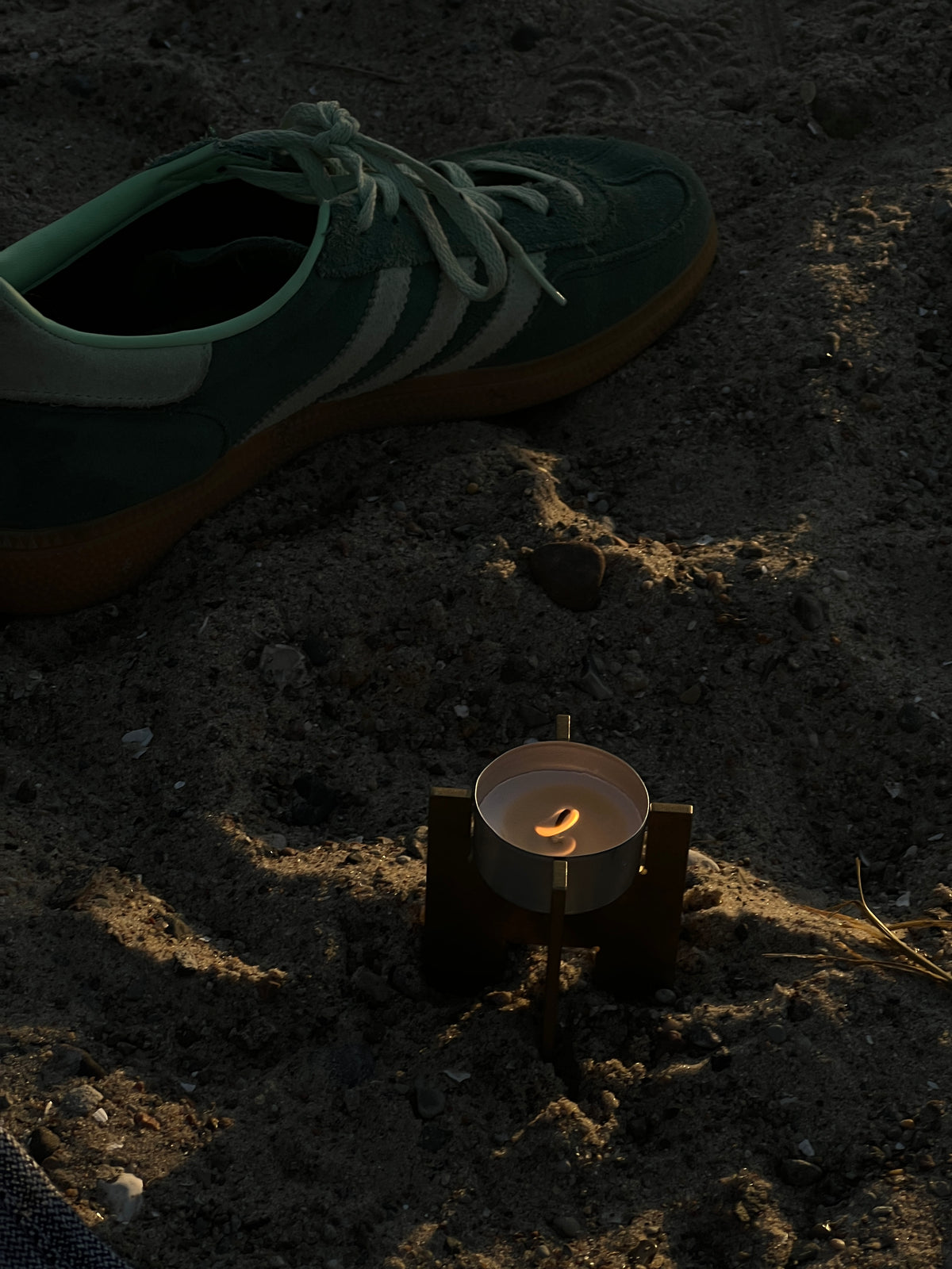 Lyssier H64 candleholder from solid brass with a lit tealight placed in the sand during golden hour, next to a pair of sneakers.