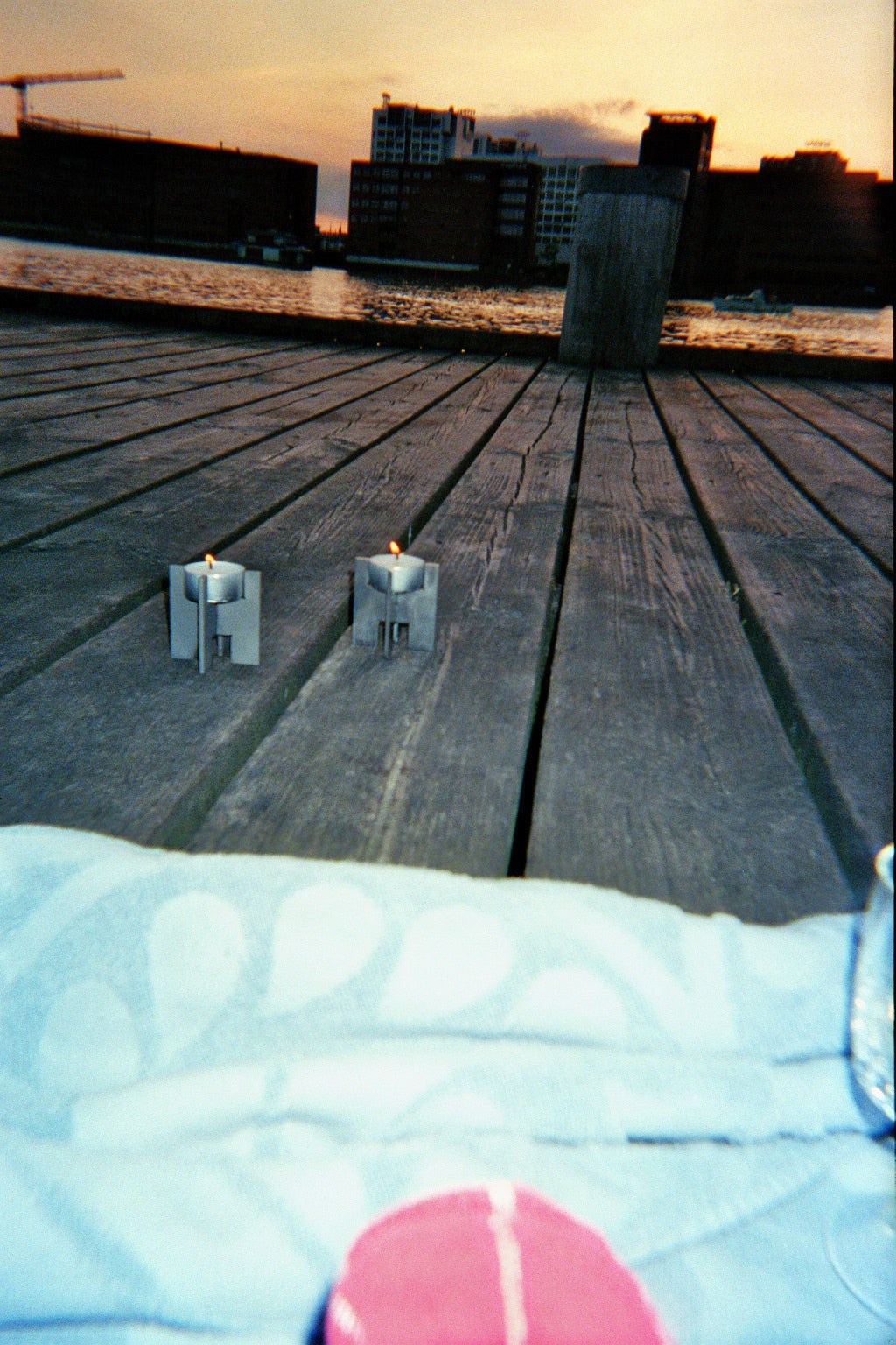 Two Lyssier H64 stainless steel candleholders with lit tealights on a wooden waterfront deck during sunset.