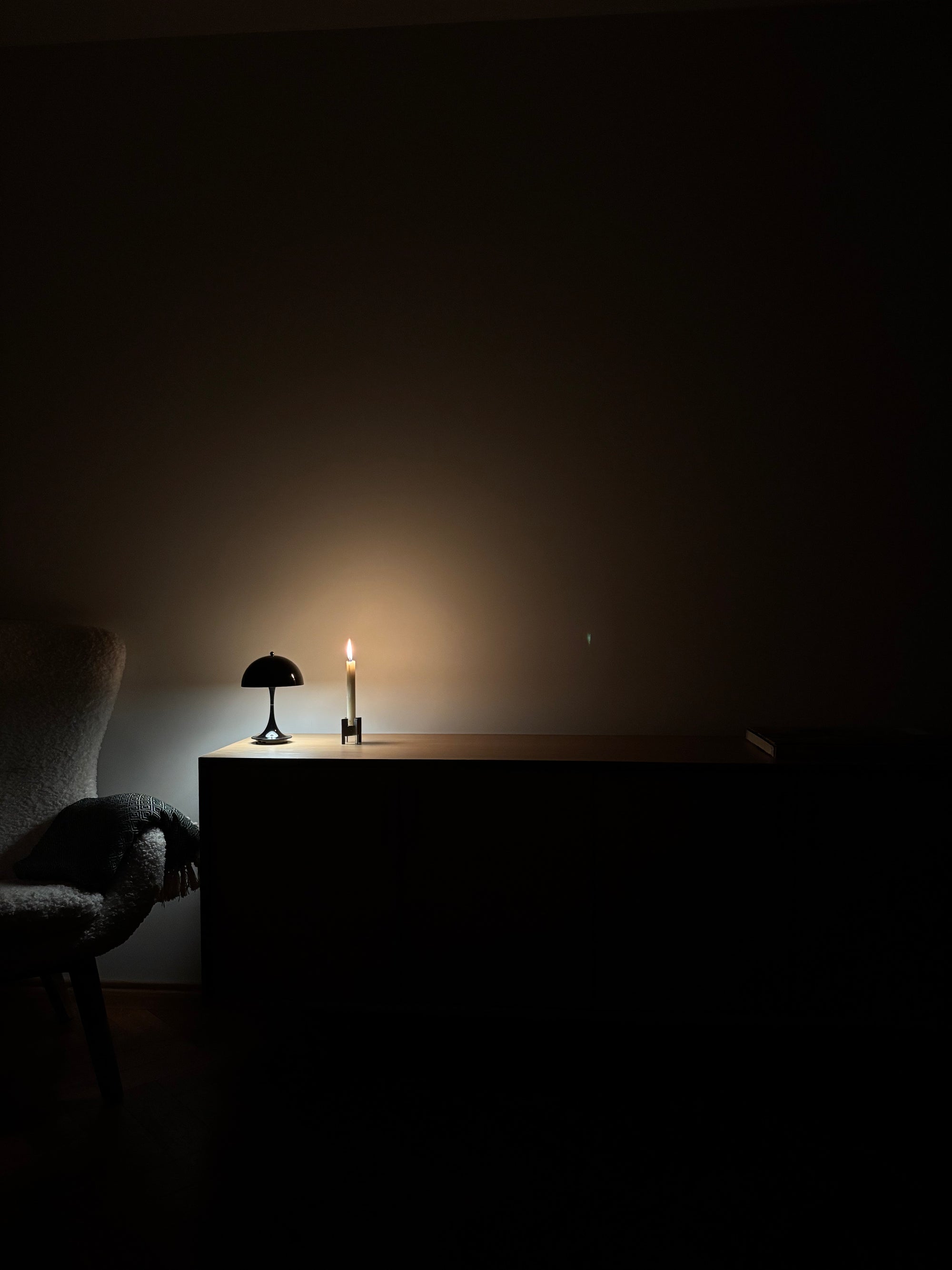 A dimly lit interior scene with a small table against a dark wall, holding a black dome lamp and a H64 candleholder with a single lit candle that casts a warm glow. Part of a chair is visible on the left.
