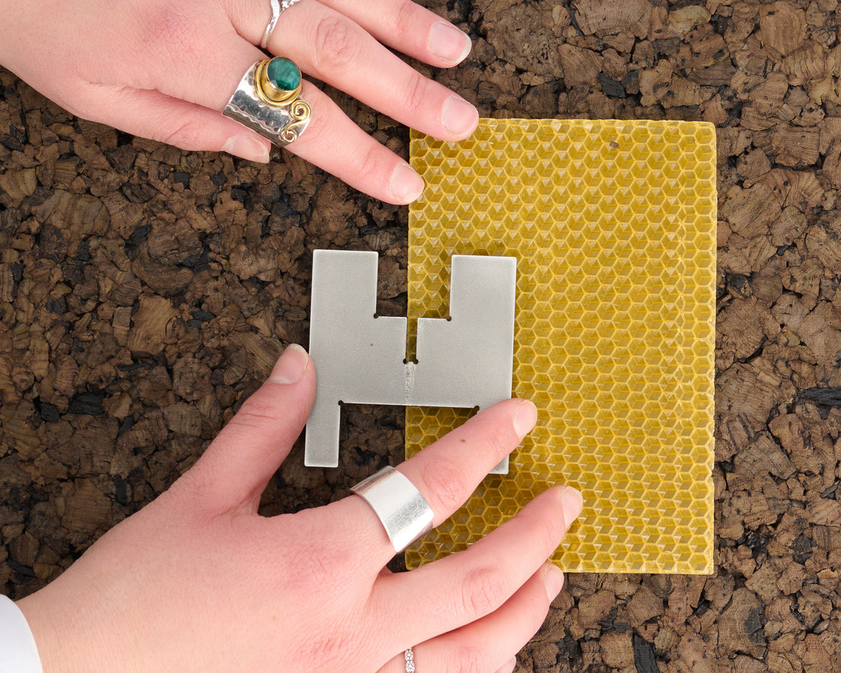 Hands beside one of the two stainless steel pieces of a Lyssier H64 candleholder, resting on a yellow beeswax sheet for rolling a portable candle on a textured cork surface.