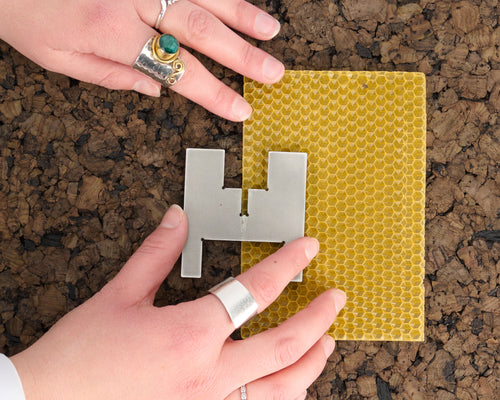 Hands beside one of the two stainless steel pieces of a Lyssier H64 candleholder, resting on a yellow beeswax sheet for rolling a portable candle on a textured cork surface.