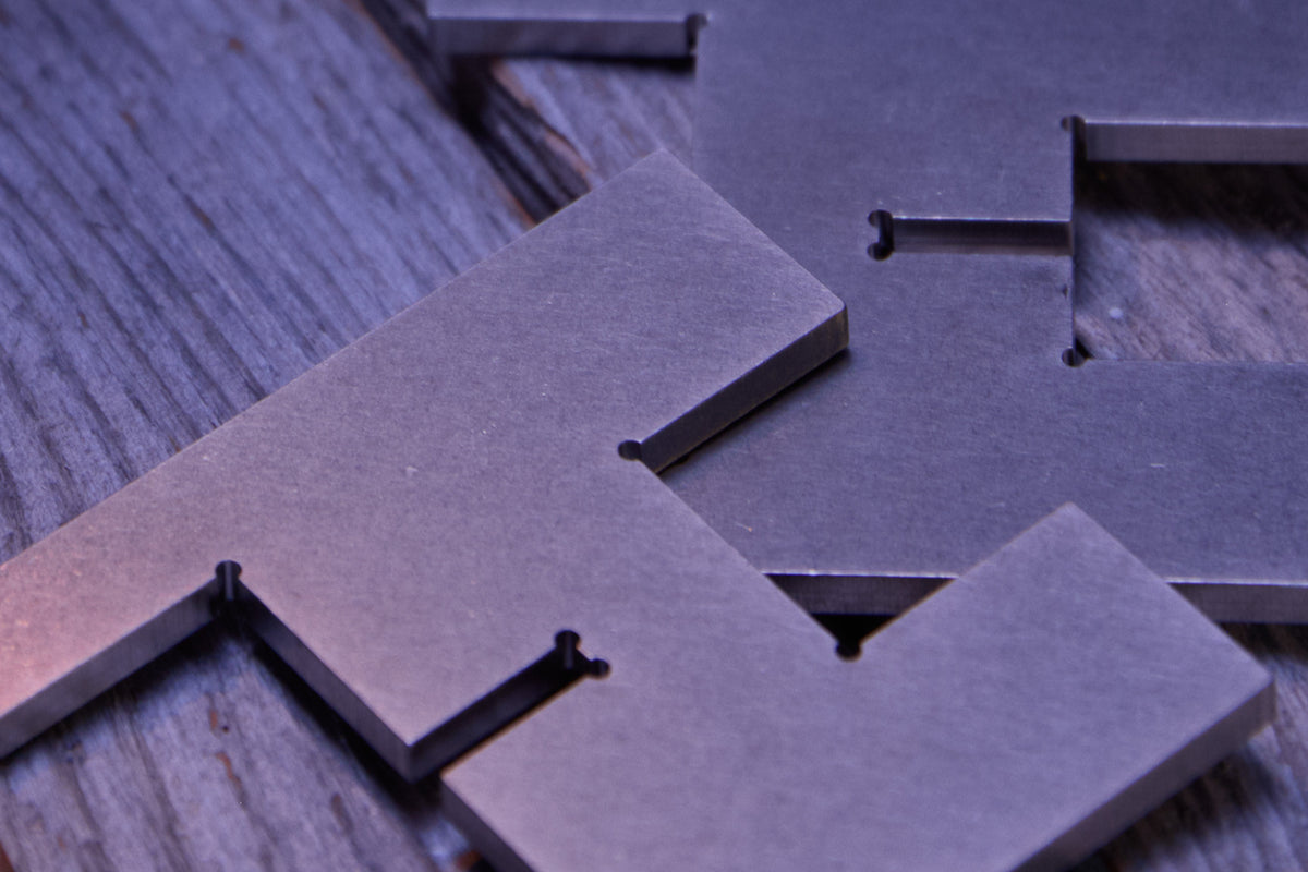 Close-up of one unassembled Lyssier H64 candleholder, showing two precision-cut solid stainless steel pieces with a matte tumbled surface and precisely formed interlocking slots, arranged on a rustic wooden table under cool blue hour light.