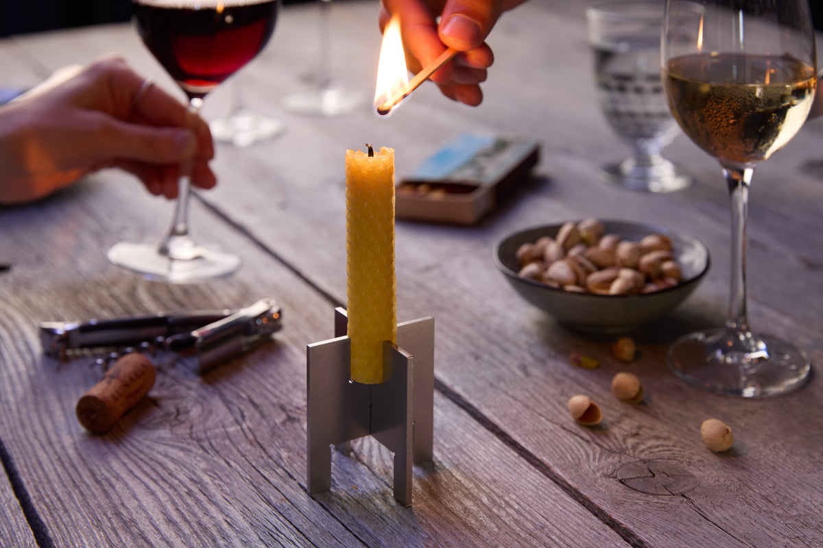 A minimalist Lyssier H64 candleholder made from two interlocking stainless steel pieces holds a yellow taper candle being lit with a match on a rustic wooden table outdoors during golden hour, surrounded by wine glasses, corks, and a bowl of nuts.