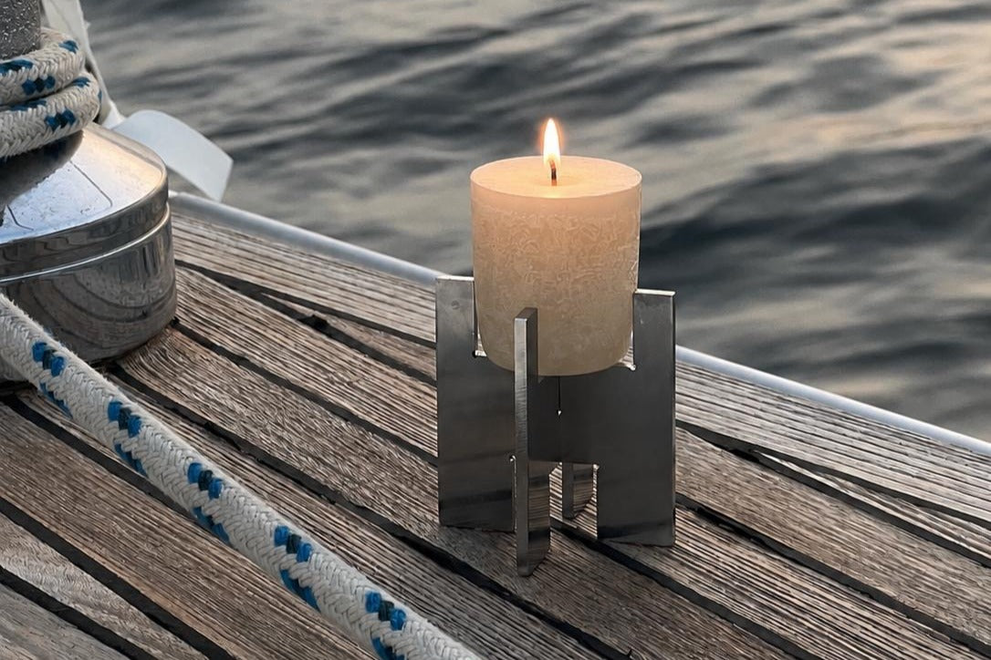 A Lyssier H64 candleholder made from two interlocking stainless steel pieces holds a lit pillar candle on the wooden deck of a sailing boat at sea.